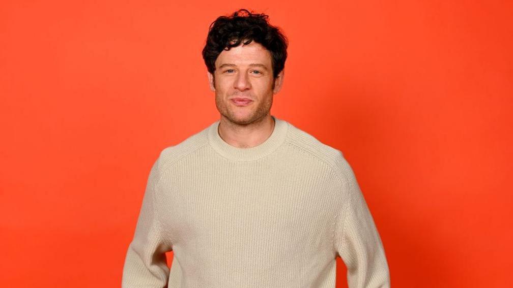 The actor James Norton wearing a white woollen jumper and standing in front of an orange back drop.