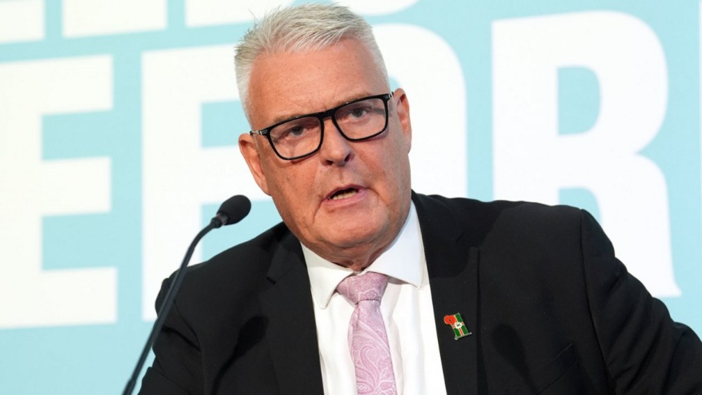 Headshot of Reform welfare spokesman, Lee Anderson, speaking at a press conference in front of a large turquoise Reform UK banner. He has short, white hair, styled with gel, and wears a black suit, white shirt, pink tie and a poppy badge.