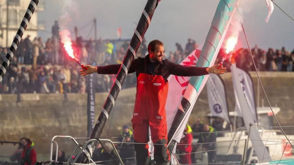 A man with a beard in a red sailing suit holds red flares in each hand. He is standing on a yacht with a crowd behind.