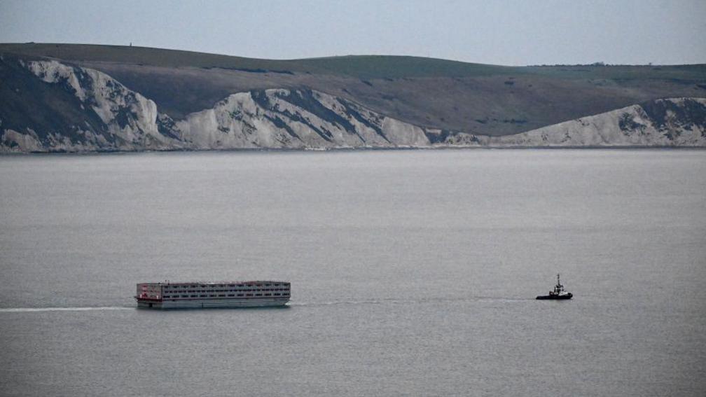 The Bibby Stockholm being towed by a tugboat, a scenic view of the coastal cliffs in the background.