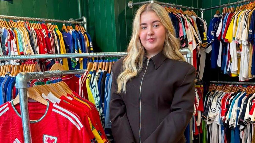 An 18-year-old girl with long blonde hair is looking directly at the camera as she's stood in her store surrounded by hundreds of old football shirts