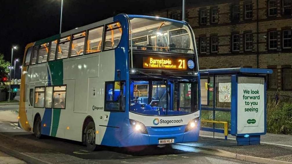 A double-decker bus, pictured at night at a deserted bus stop. Operated by Stagecoach, the bus is painted blue, white, green and yellow and has  "Barnstaple" on its destination board.