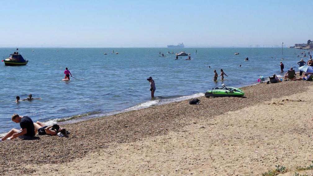 People sunbathe along the shingle on a beach, with lots of people in the water. There are also some boats. It is a sunny, clear day.