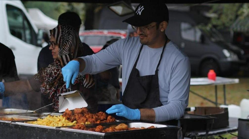 Man preparing chips at the Middlesbrough Mela. He is wearing gloves, a grey shirt, black apron and a black cap. A woman next to him is also cooking. 