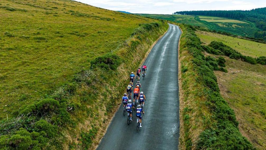 An aerial view of a group of cyclists on a narrow country road in the hills. The road winds round a bend in the distance with green slopes on either side.