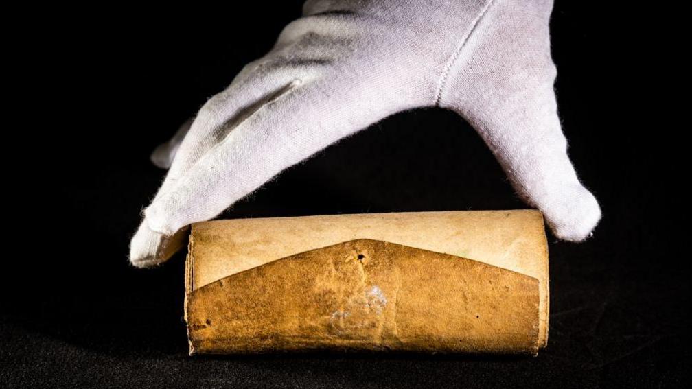 Rolled up brown paper on a black background. It is being held by a white glove.