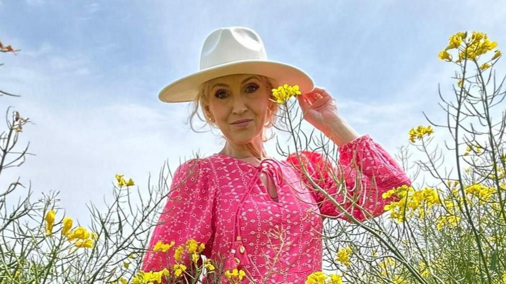 Yn y llun yma mae Rhian yn yr ardd, wedi ei hamgylchynu gan flodau melyn. Mae hi'n gwisgo ffrog binc gyda hat gwyn.