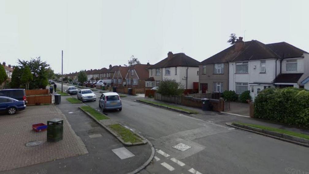 A screenshot from Google street view showing a residential street with semi-detached houses on one side and a short row of shops on the other. There are cars parked on one side of the street and it's a cloudy day.