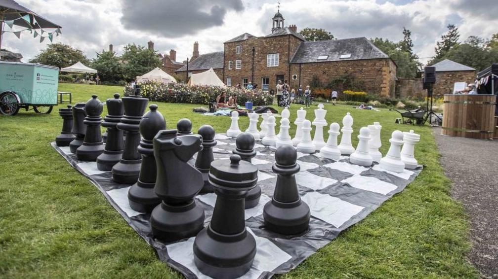 Stately home in background, a large chess set on grass in the foreground