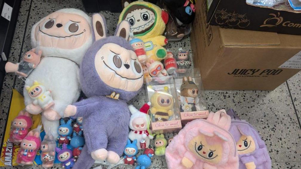 Dolls of different sizes and colours are on the floor. Two cardboard boxes are on the top right of the photo.