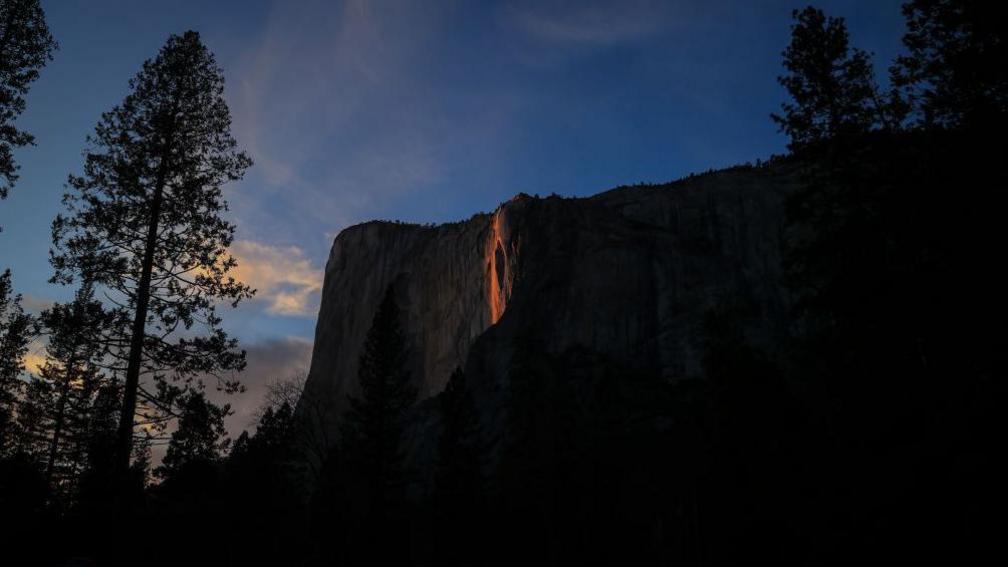 view of the side of El Capitan mountain, California with a waterfall glowing orange with the setting sunlight
