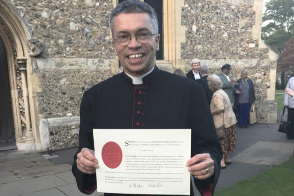 David Tudor holding up a certificate and smiling at the camera. He has thin-rimmed glasses on and is wearing black clerical clothing.