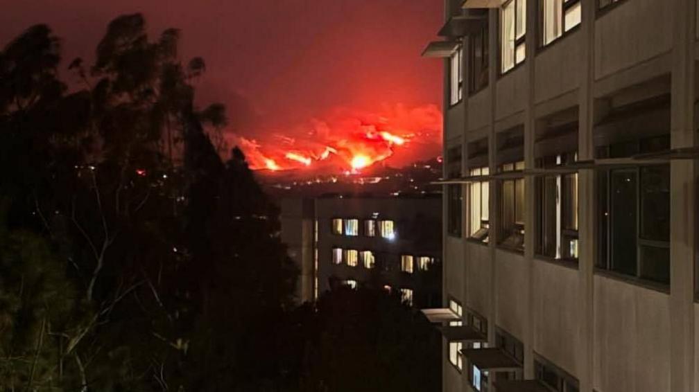 An image of the LA wildfires taken from the UCLA campus. It is night time and in the distance the landscape is burning red. A dorm block with lights on can be seen in the foreground.