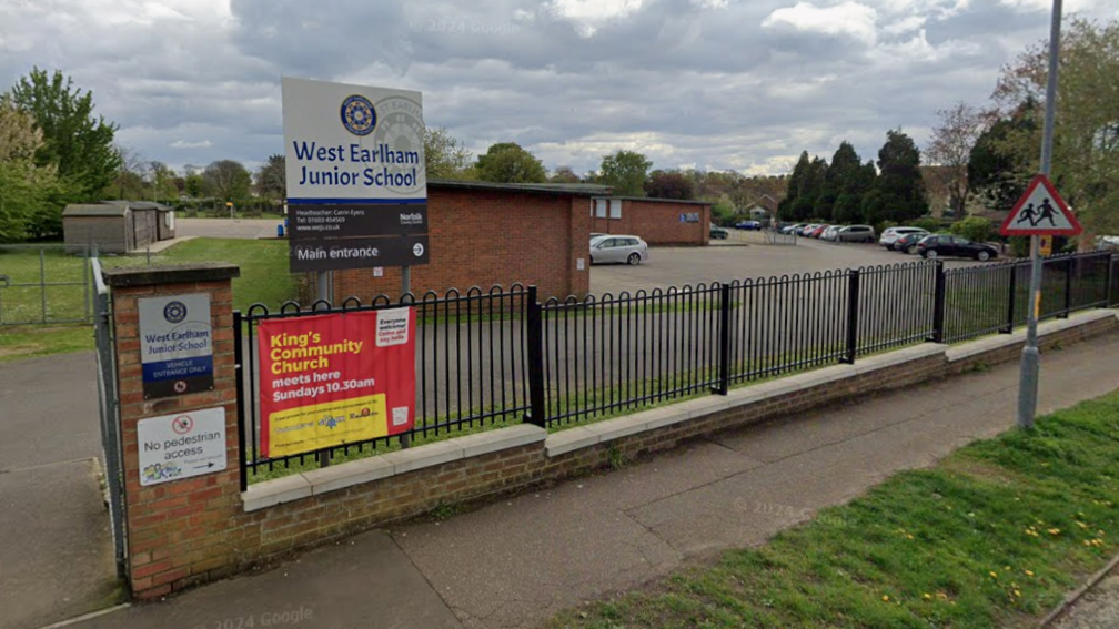 The outside of a school. There are gates outside the front of the building and a number of signs. There are cars in the car park.
