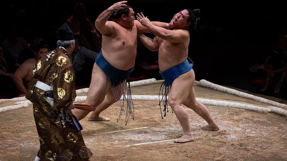 Sumo wrestlers Tamawashi (left) battles with Wakatakakage (right) at the centre of the dohyo, watched by a gyoji (sumo referee)