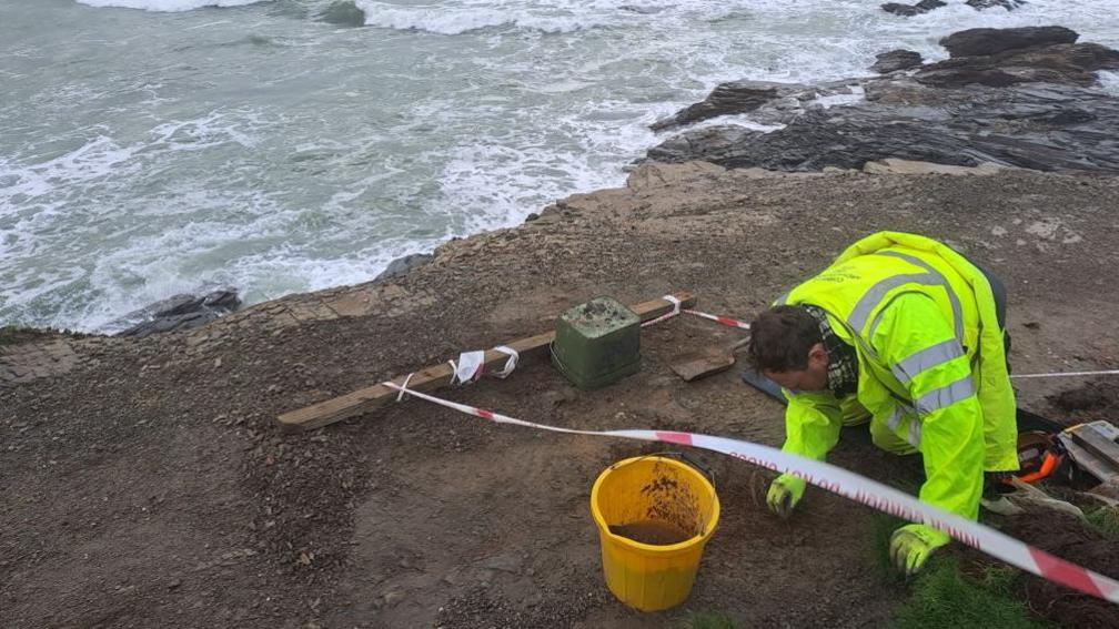 A man looking at the soil along the north Cornish coast. He is metres away from the sea and the man has a yellow high-vis jacket on and a yellow bucket next to him.