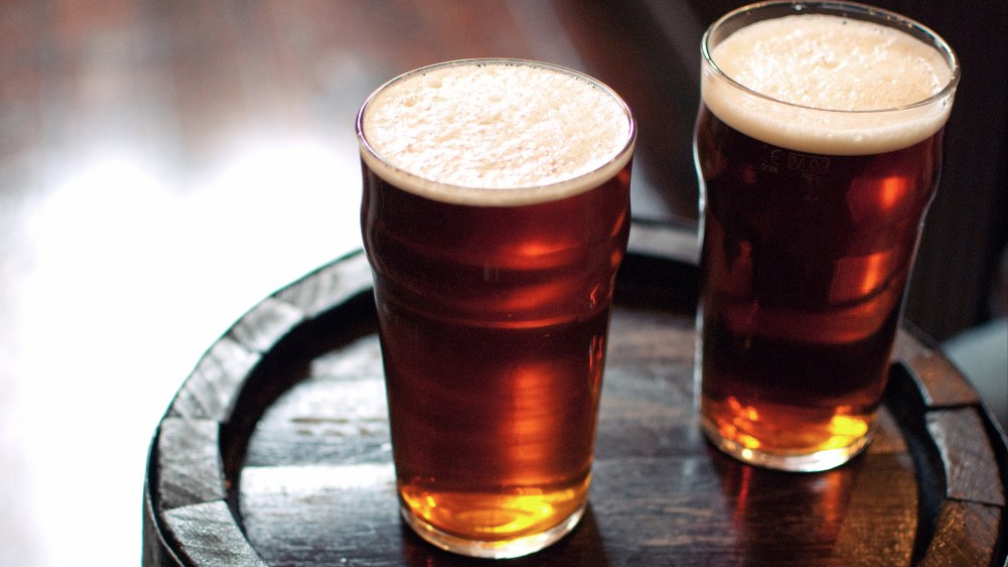 Two pints of beer on a small table which appears to be a beer barrel. The drinks have small heads.