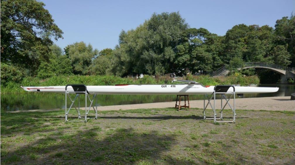 Image shows the boat on a stand near the river and a bridge

