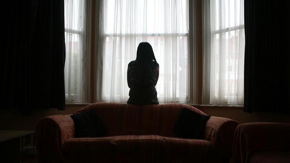 A woman sits on the back of a sofa, looking out of the window in fear of her partner, coming home after football.