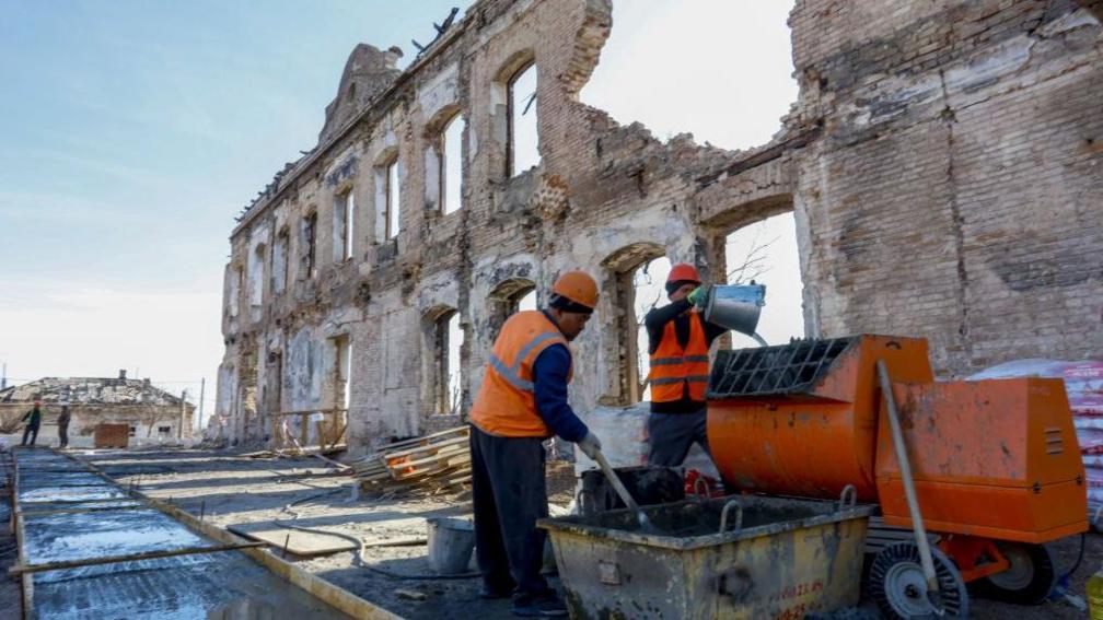Russian workers in Mariupol mix building materials. They are dressed in hard hats and orange high-vis jackets. Behind them is a war-damaged building with a chunk of its wall missing.
