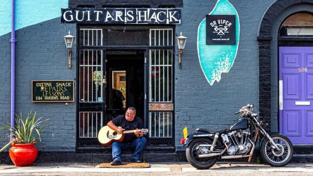 A man sits on a sunny day playing  an acoustic guitar in the street