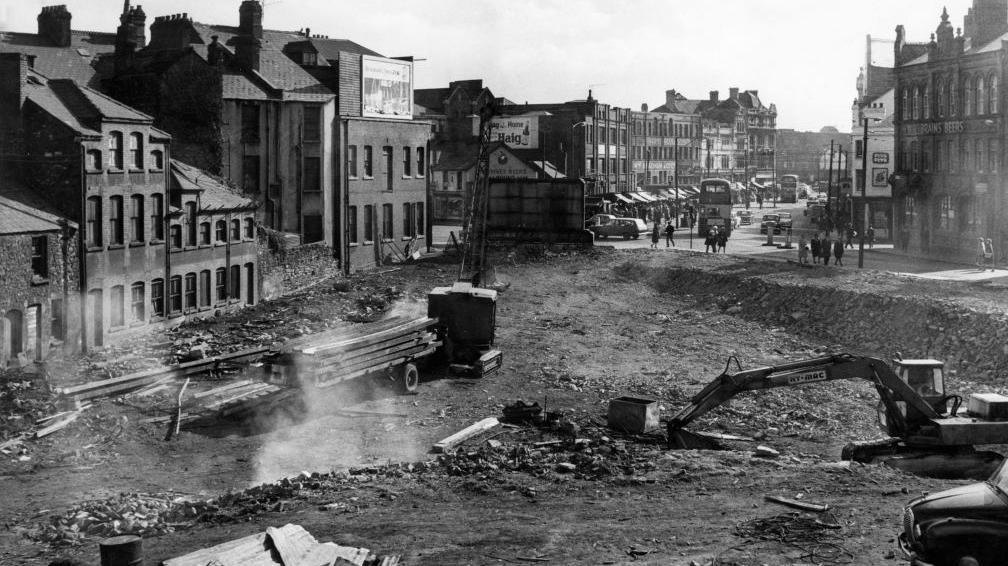 A black and white photo of The Hayes in Cardiff under construction, with construction vehicles in a large working area, and buses, cars and people in the background