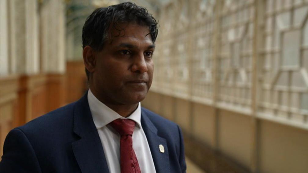 Majid Mahmood has dark hair and is wearing a navy suit jacket, white shirt and red tie