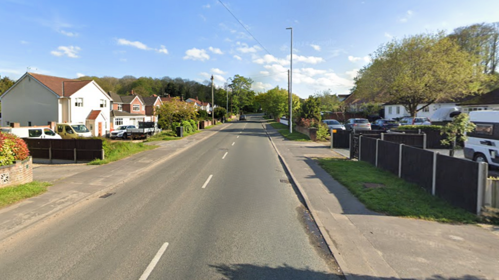 Houses lining either side of a stretch of road. A number of them have cars parked in their drives. 