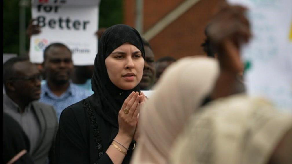 A woman dressed in a black hijab in a crowd holding her hands up in prayer