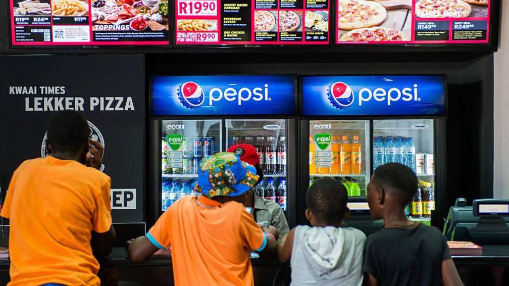 Four boys, two in orange t-shirts, stand at a pizza fast food counter ordering food with two drinks machines advertising Pepsi in the background