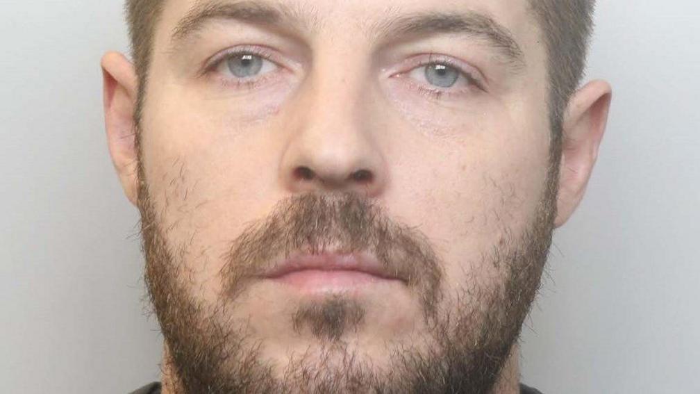 A police photograph of Robin Vaughan, he looks at the camera with a neutral expression. He has short brown hair and a short brown beard