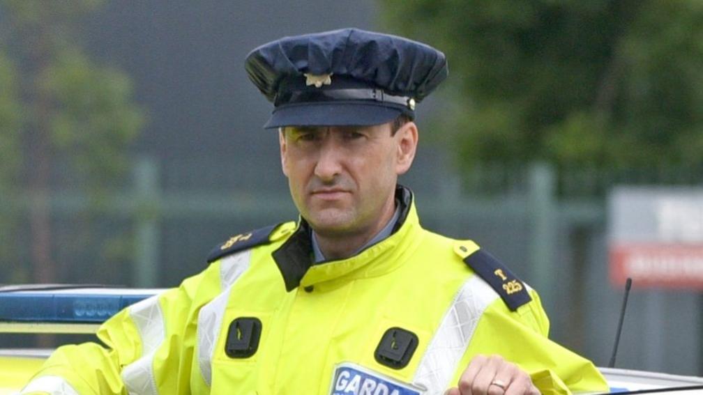 A man wearing an Irish police officer uniform of a yellow hi-vis jacket standing behind the open door of a police car which is stationary.