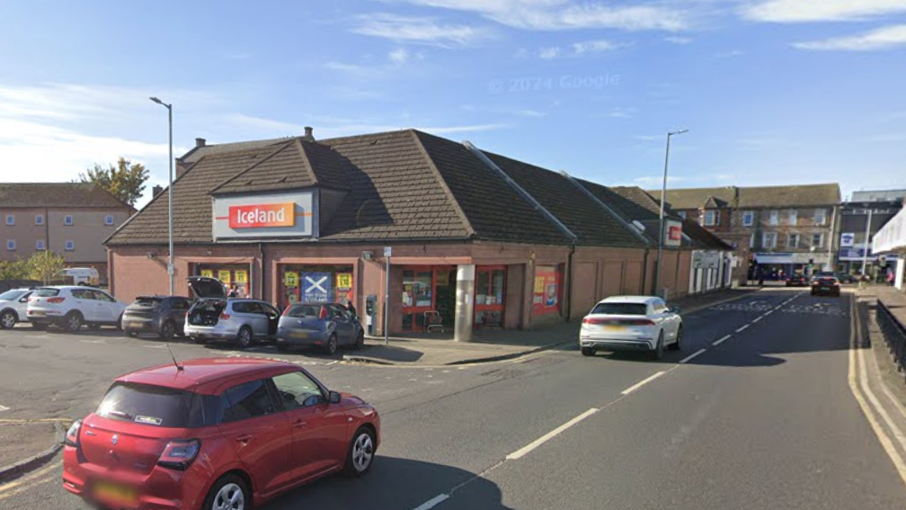 Fowlds St in Kilmarnock, shows a red and white car driving down the street an Iceland supermarket is to the left and a car park is on the right.