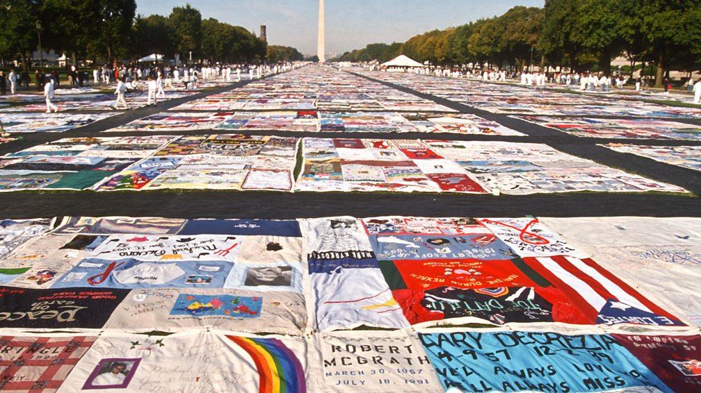 Many quilts with text and images on them on display in a flat outdoor space on a sunny day