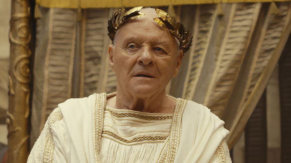 Anthony Hopkins dressed in white and gold emperor clothes stands in front of a curtain with soldiers to his left