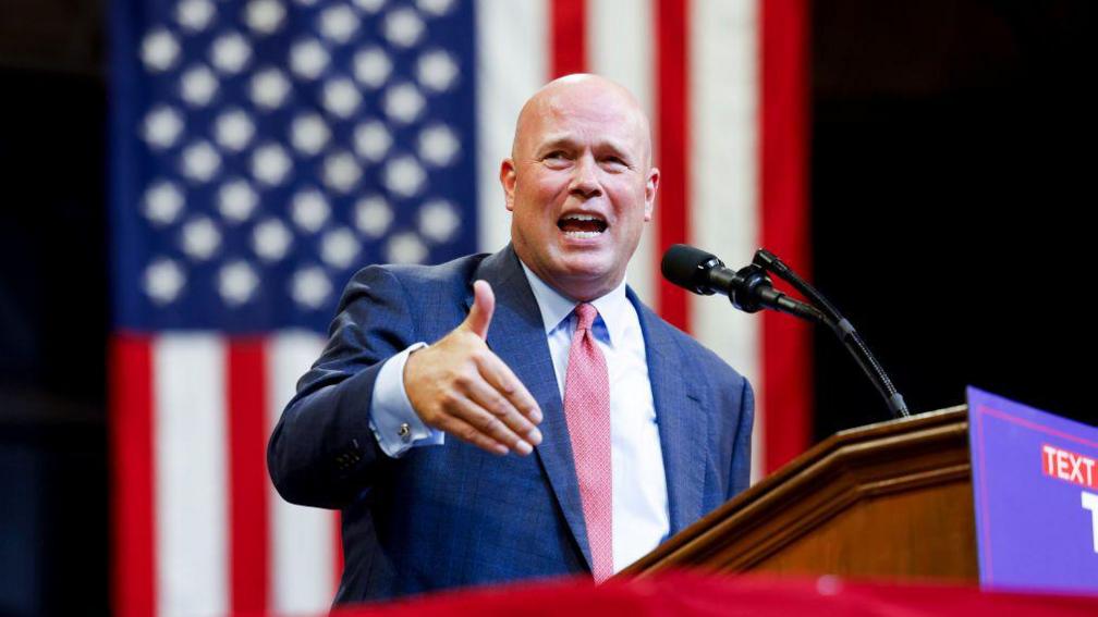 Matthew Whitaker makes a speech in front of a lectern while gesturing with his hand. He is bald, wears a navy suit, white shirt and pink polka dot tie. There is an American flag hanging behind him.