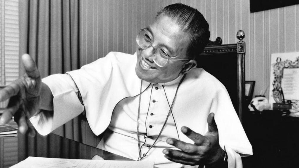 Cardinal Jaime Sin is seen in his office on 26 February, 1986 - he is sitting behind a desk and smiling as he gestures.