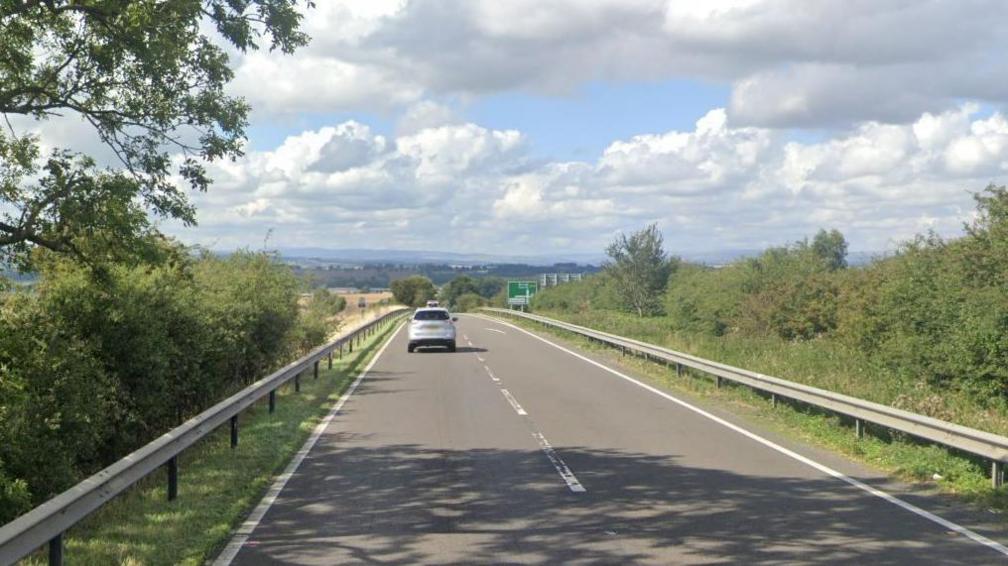 A dual carriageway road with trees on either side and a view of hills in the distance. A silver car on the road. Blue sky with clouds.