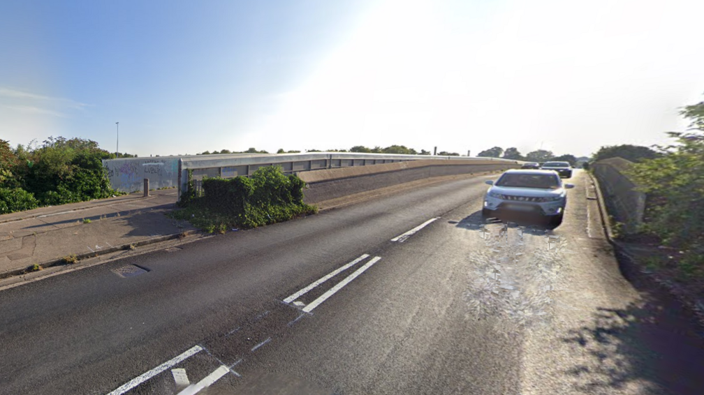 A Google streetview image of the railway bridge on Westfield Road near Bourges Boulevard in Peterborough. At least two cars are driving towards the camera. There are blue skies with some white cloud.