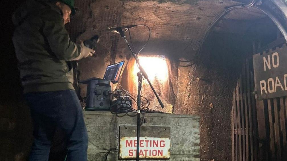 Liam Gaughan recording the sound at Mahogany Drift Mine. He wears jeans, a green jacket and a helmet. He is holding a device near a standing microphone, with laptop and other recording equipment near him. There is a sign reading Meeting Station and another on a wood-panel door reading No Road.