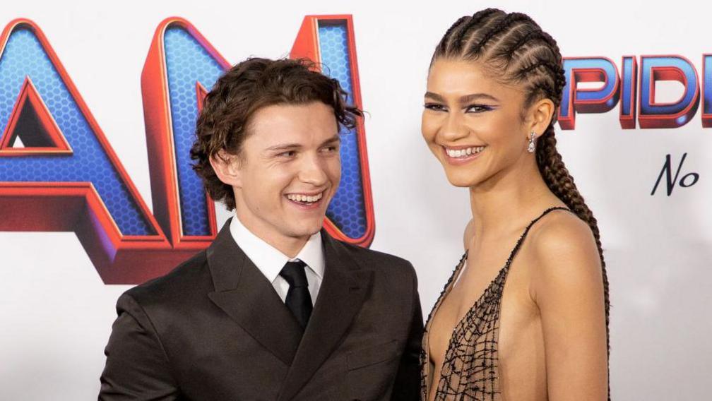 Actor Tom Holland wearing a brown suit and Zendaya wearing a black patterned dress