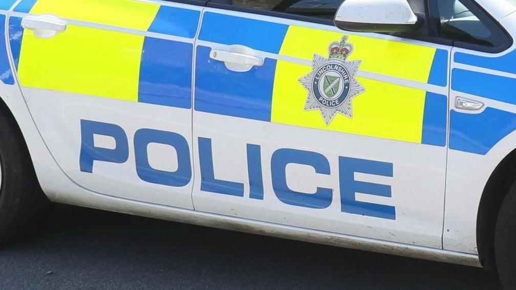 A close-up of the side of a police car, which is coloured white with blue and yellow panels, a Lincolnshire Police logo and the word "police" written in large capital letters. 