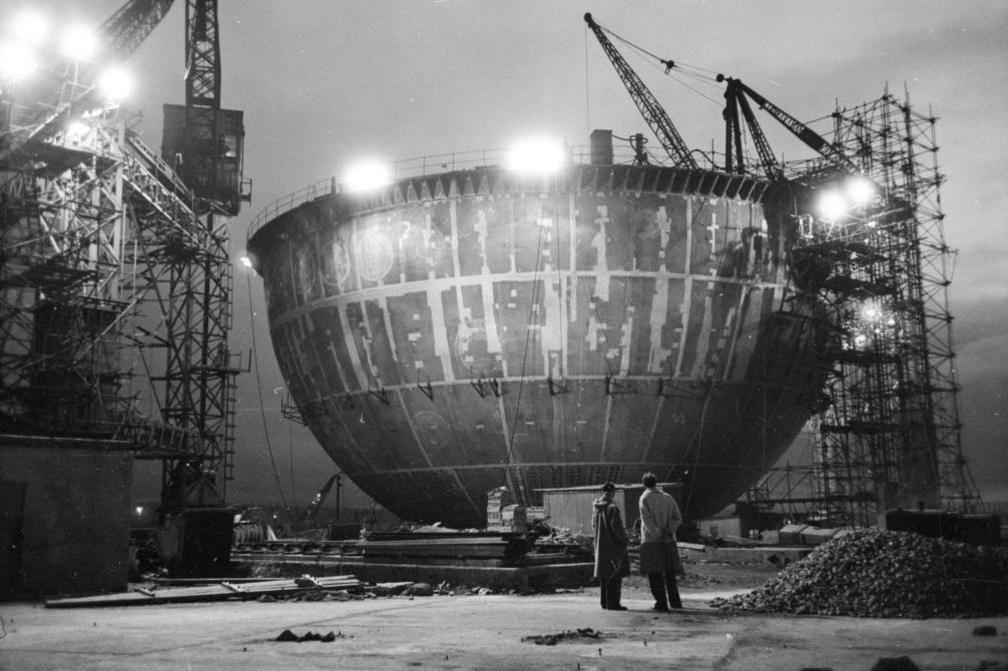 In the black and white image two men look up at the partly-completed steel dome. There is scaffolding, a crane and floodlights.