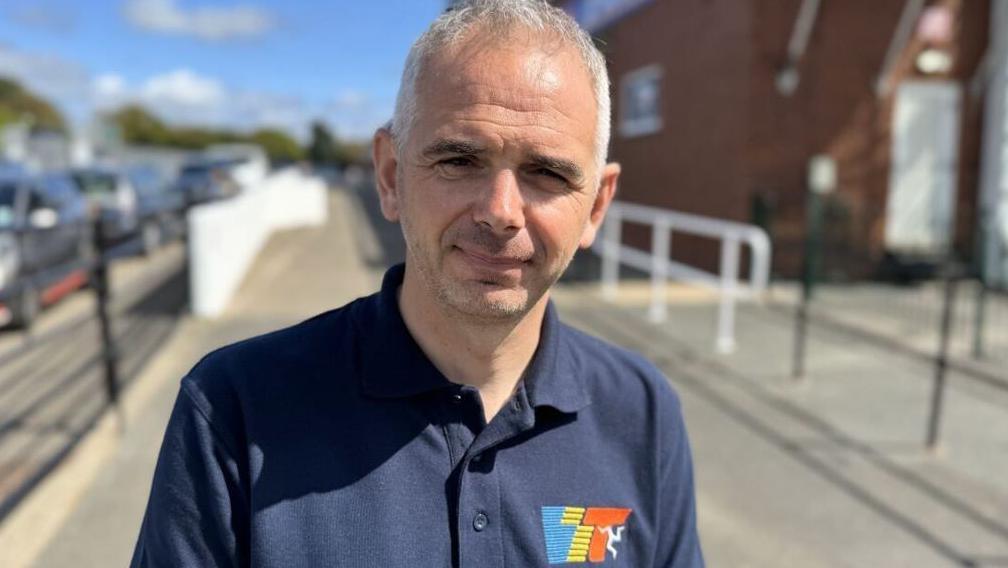 Paul Phillips wears a blue polo shirt with a colourful logo. He has short grey hair.