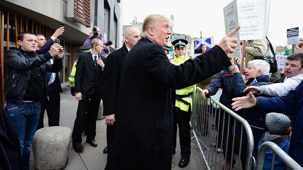 A photo from 2012 of Donald Trump outside the Scottish Parliament at Holyrood in Edinburgh. Behind a barrier are protesters and some people who appear to want to shake his hand. Police and photographers are behind Trump as he talks at the crowd with his hand raised and his finger pointed. 