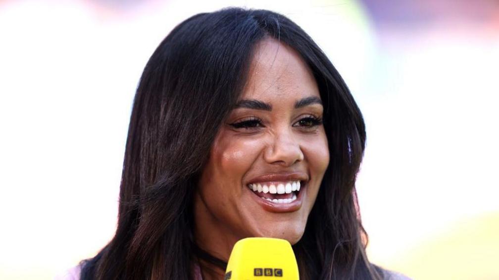 Alex Scott laughs as she presents on the BBC, she has her long black hair down