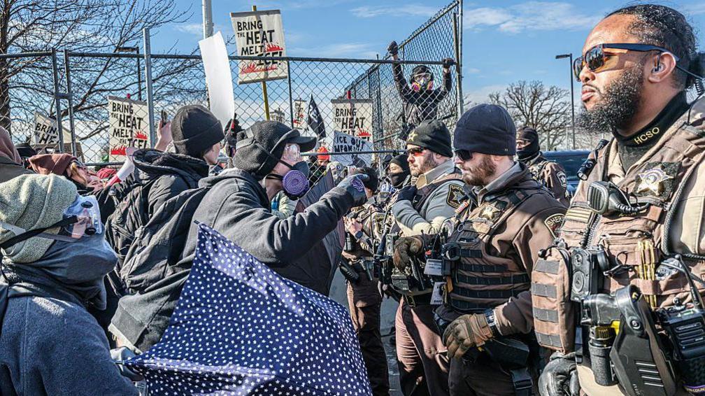 Law enforcement officers standing in a line in front of protestors wearing gas masks
