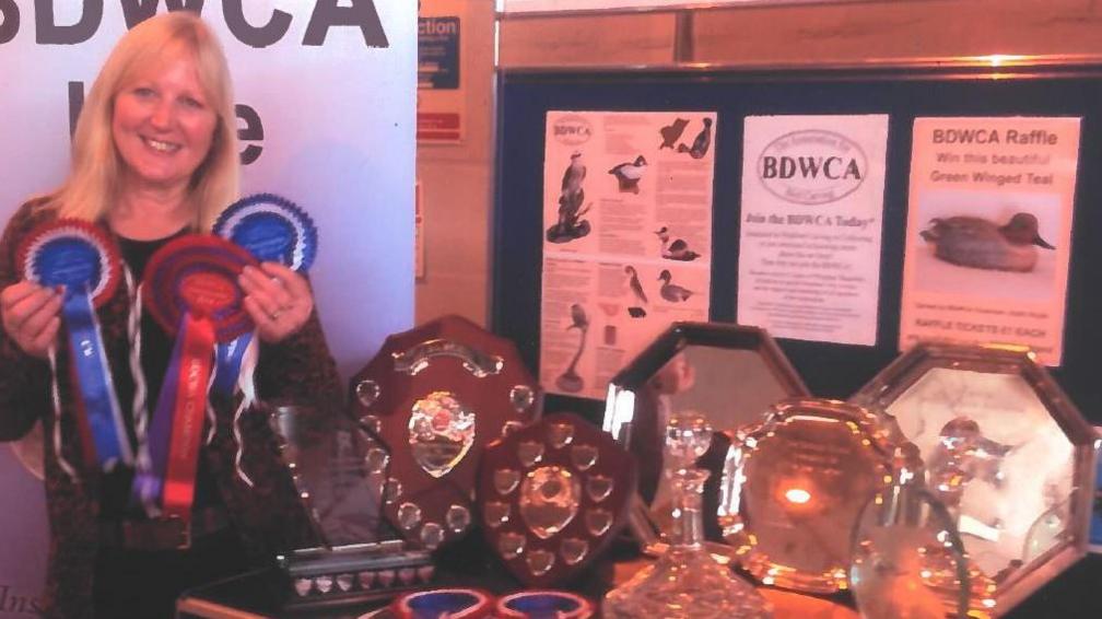 A photograph of Maggie Port, who is in the left hand side of the frame, holding several prize ribbons. To the right of her is a table full of shields and trophies. Behind the trophies is a cork board with various pamphlets from the carving club pinned to it.