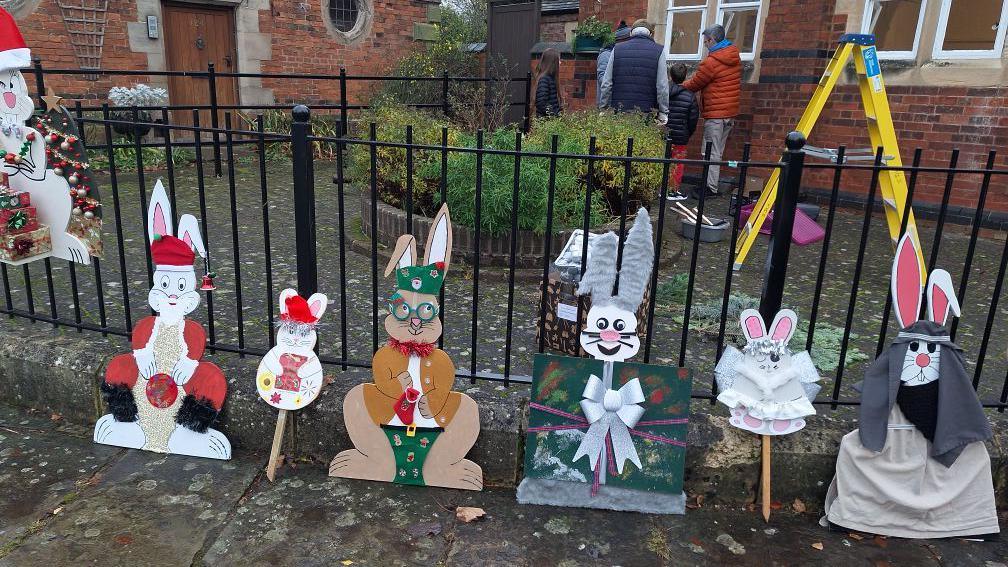 Five large and two small rabbit figures, decorated as Christmas characters, leaning against a set of railings