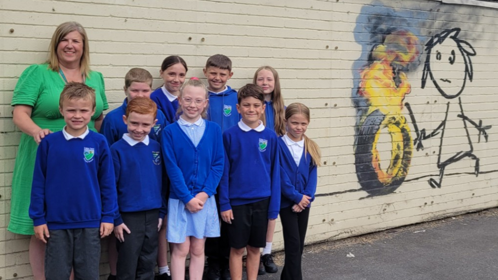 Children from Bridge Farm Primary school in Bristol smile at the camera. They are wearing blue jumpers and standing against a wall which has a piece of Banksy art on it - a child rolling a burning tyre